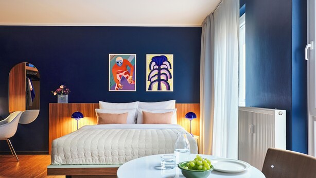 Una habitación de hotel moderna con una cama grande, una mesa de comedor redonda con un cuenco de fruta y dos cuadros abstractos en la pared.