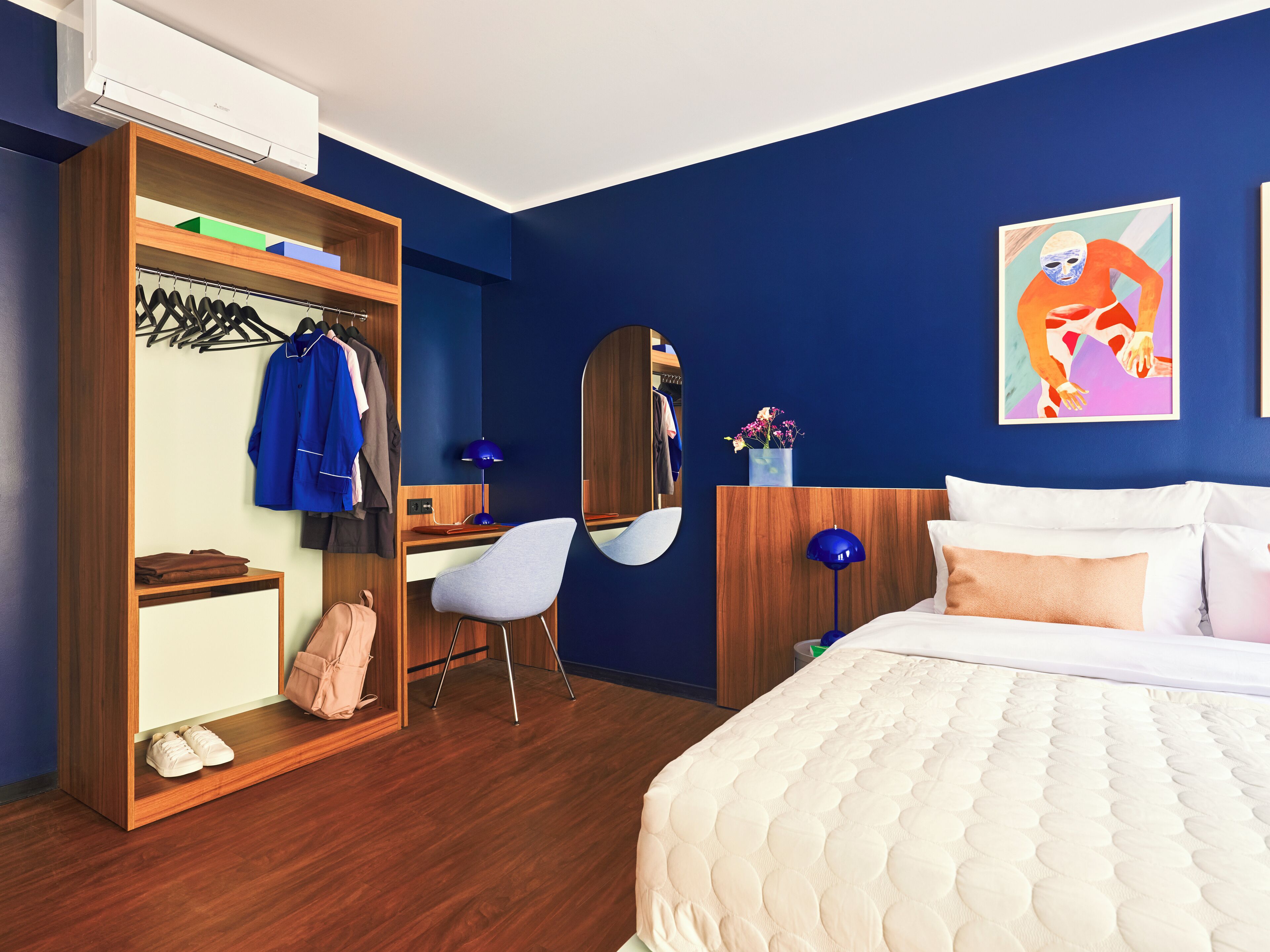 Une chambre d’hôtel avec une armoire en bois, un bureau avec une chaise, un lit avec du linge de lit blanc et un mur bleu orné d’un tableau.