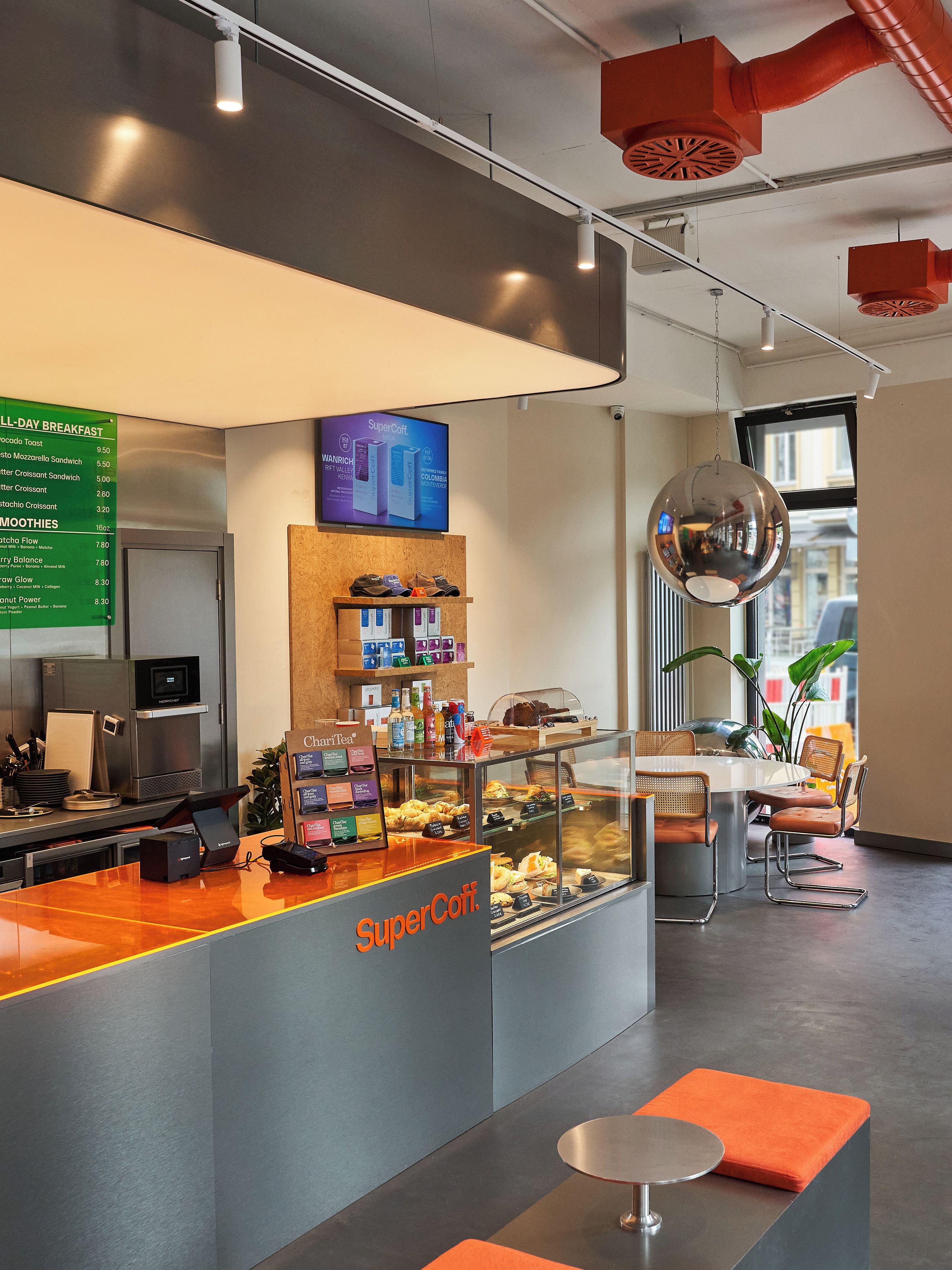 Un área de desayuno moderna con un menú digital, una estación de café y asientos con cojines naranja.