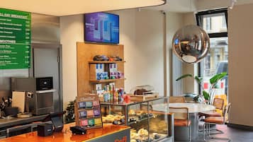 A modern breakfast area with a digital menu board, a coffee station, and seating with orange cushions.