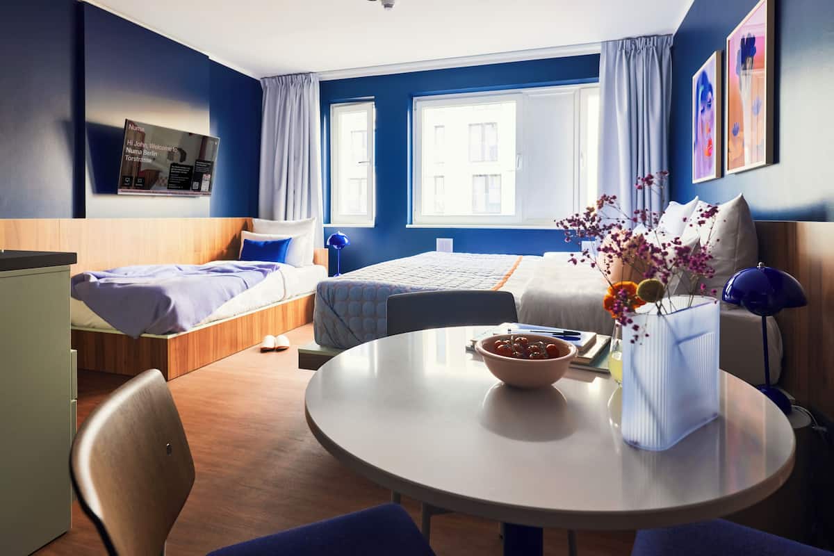 A hotel room with two beds, a dining table, a TV, and a window with curtains.