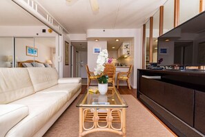 A modern hotel room with a sofa, a glass-top coffee table with a vase of flowers, wicker chairs, a kitchen area, and a TV on a wooden cabinet.