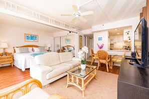 A spacious living room with a large sofa, a coffee table, and a dining area with wicker chairs. A ceiling fan is visible, and there are framed pictures on the walls.
