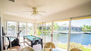 A pool table, exercise equipment, and a dining table with chairs in a room with large windows overlooking a body of water. -  Waterfront House | Private Heated Pool &. Jacuzzi| 1 mile to beach| 5 Beds (Marco Island)