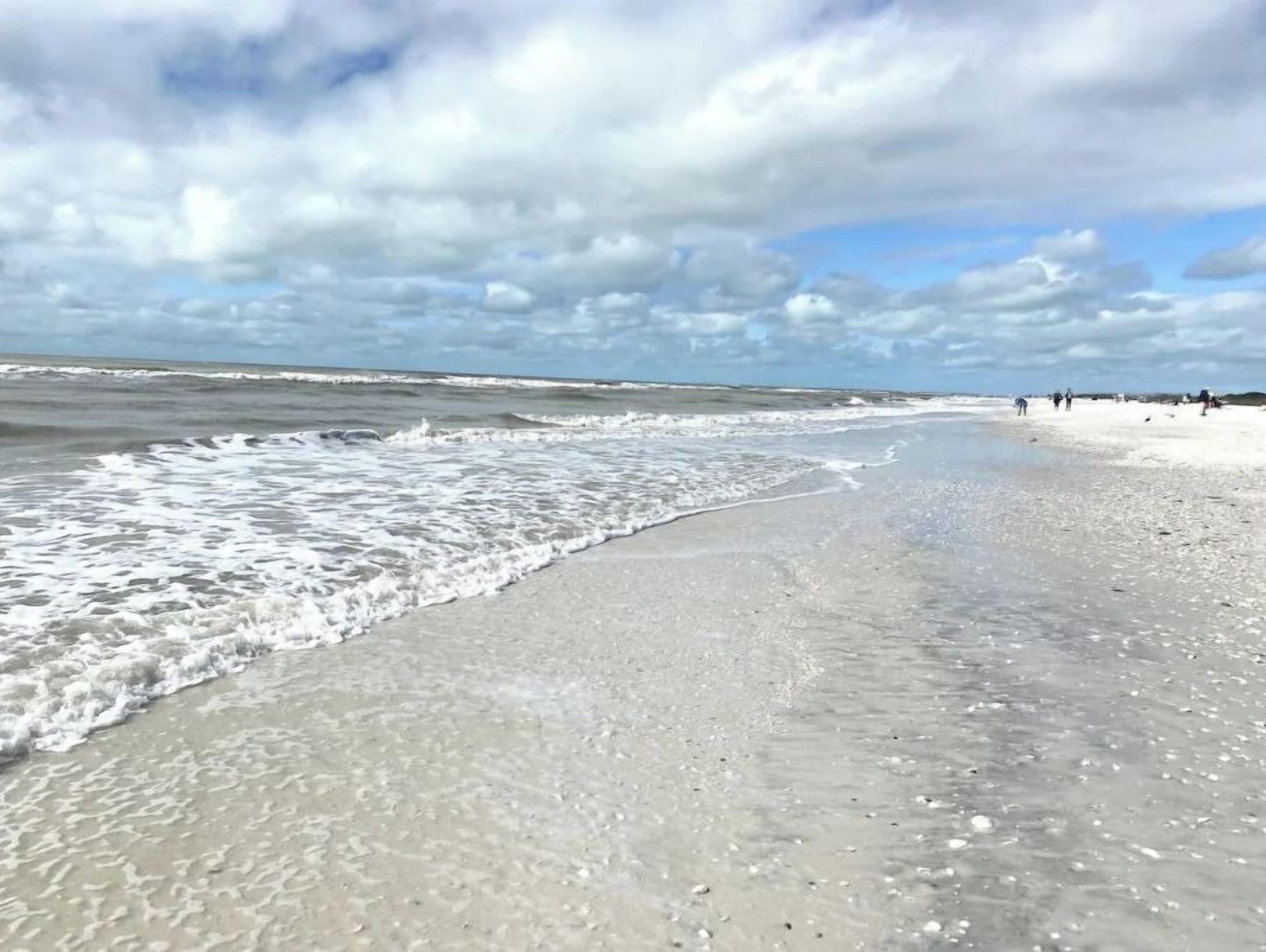 A beach with waves gently lapping onto the shore, a clear sky, and a few people in the distance.