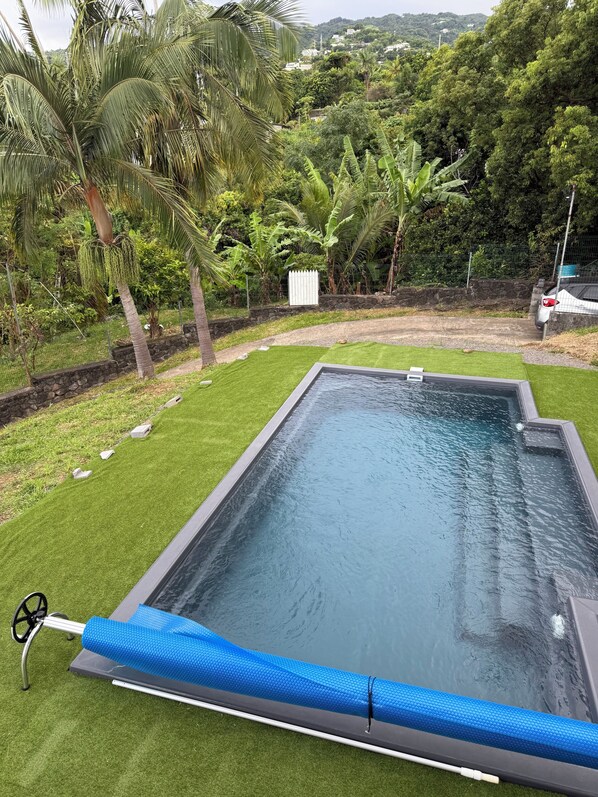 A rectangular swimming pool with a blue safety mat, surrounded by green artificial grass and palm trees.