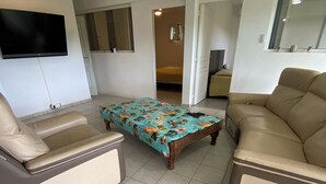 Un salon avec un canapé beige, un fauteuil assorti, une table basse en bois recouverte d’une nappe à motifs floraux et un téléviseur à écran plat fixé au mur.