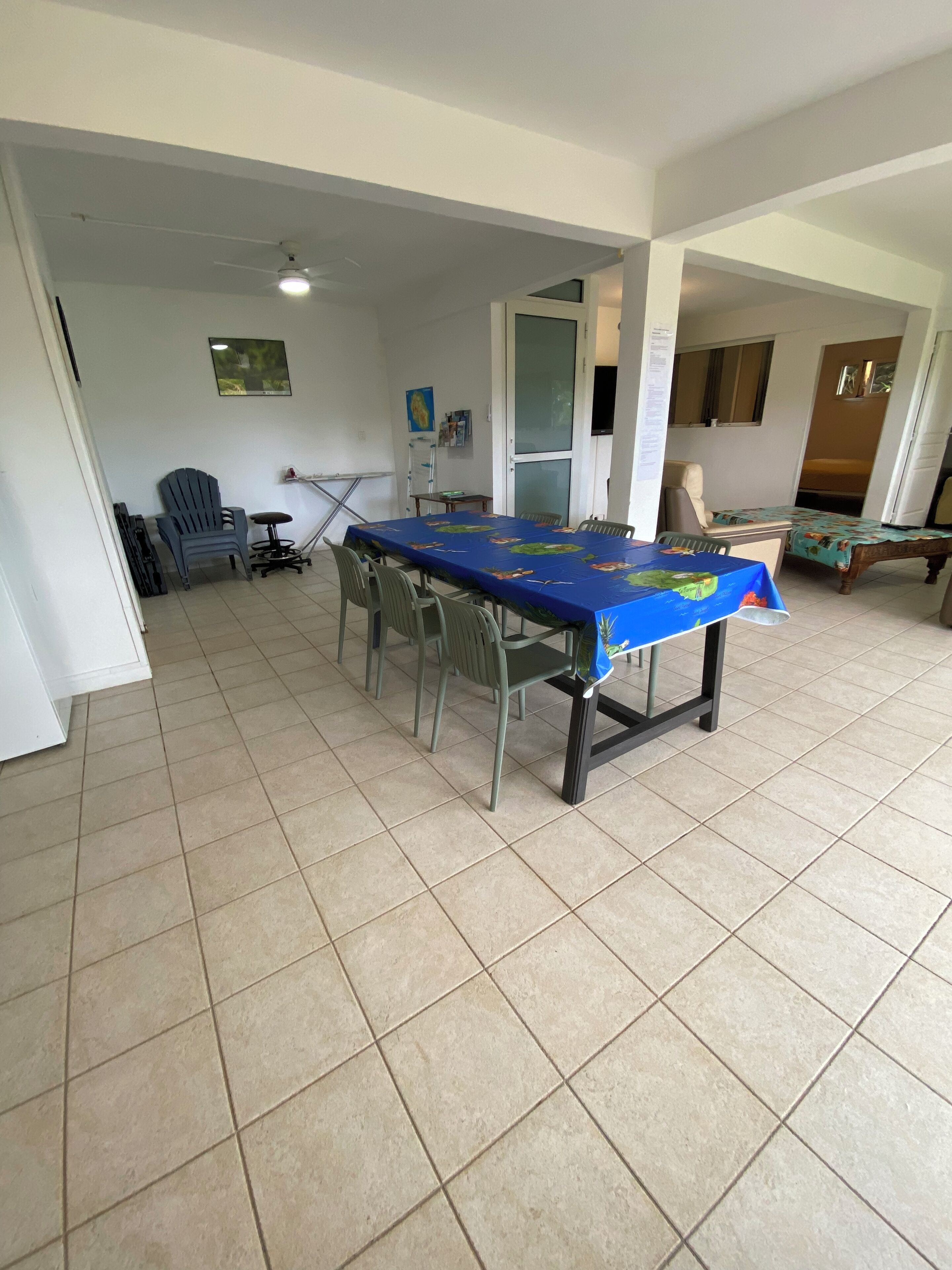A spacious room with a large dining table, several chairs, a blue plastic chair, a bed with a floral bedspread, and a ceiling fan.