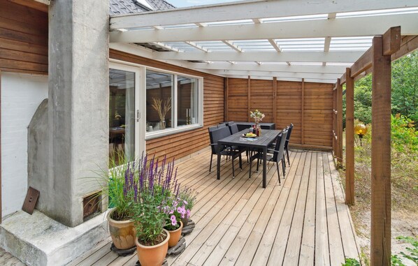 A wooden deck with a table and chairs, potted plants, and a pergola with a glass roof.