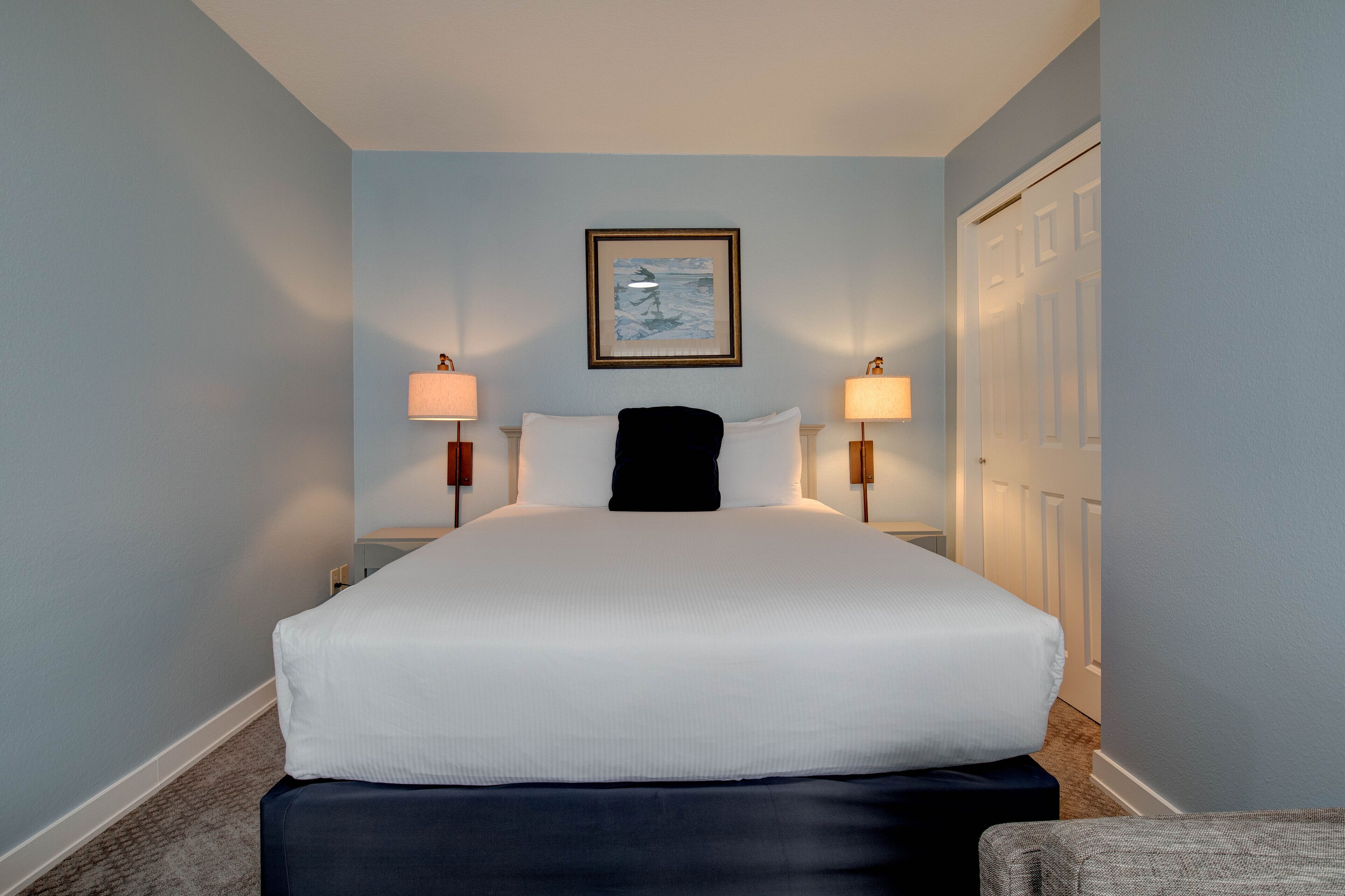 A hotel room with a large bed, two bedside lamps, a framed picture on the wall, and a door to the right.