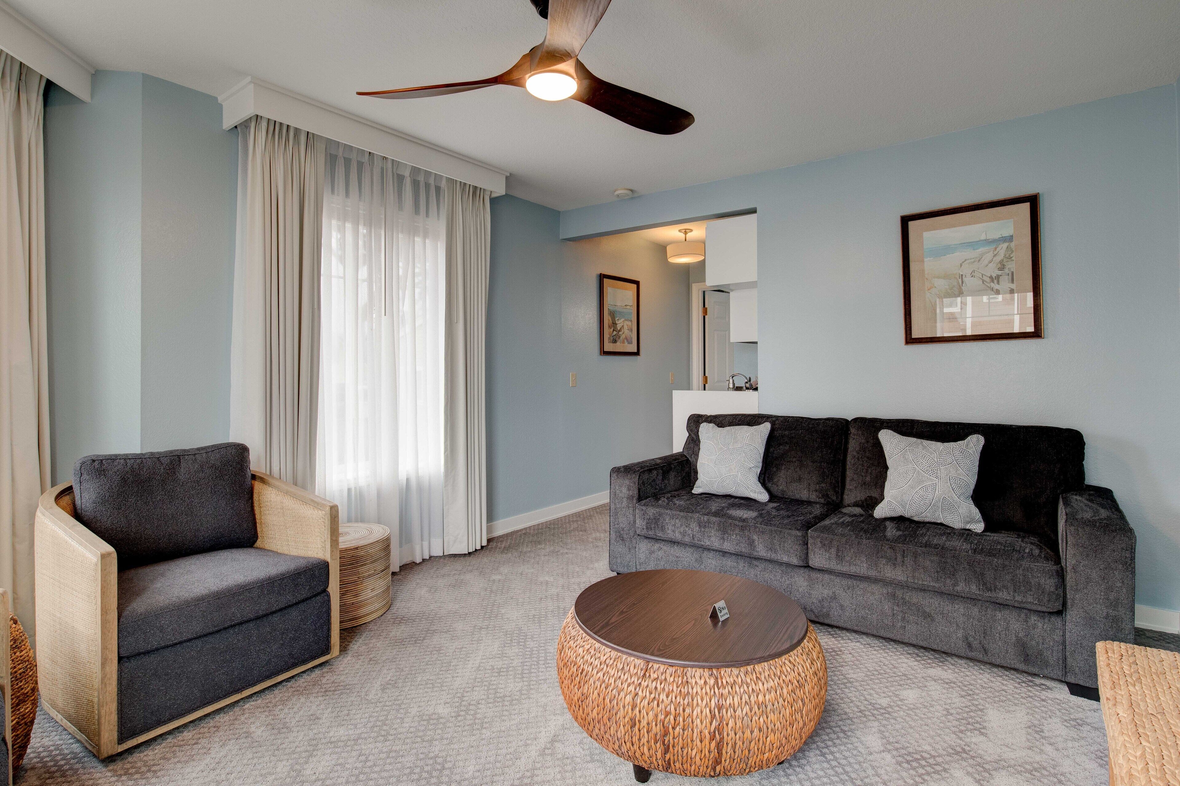 Un salon avec un canapé gris foncé, un fauteuil gris clair, une table basse ronde en osier et un ventilateur de plafond.