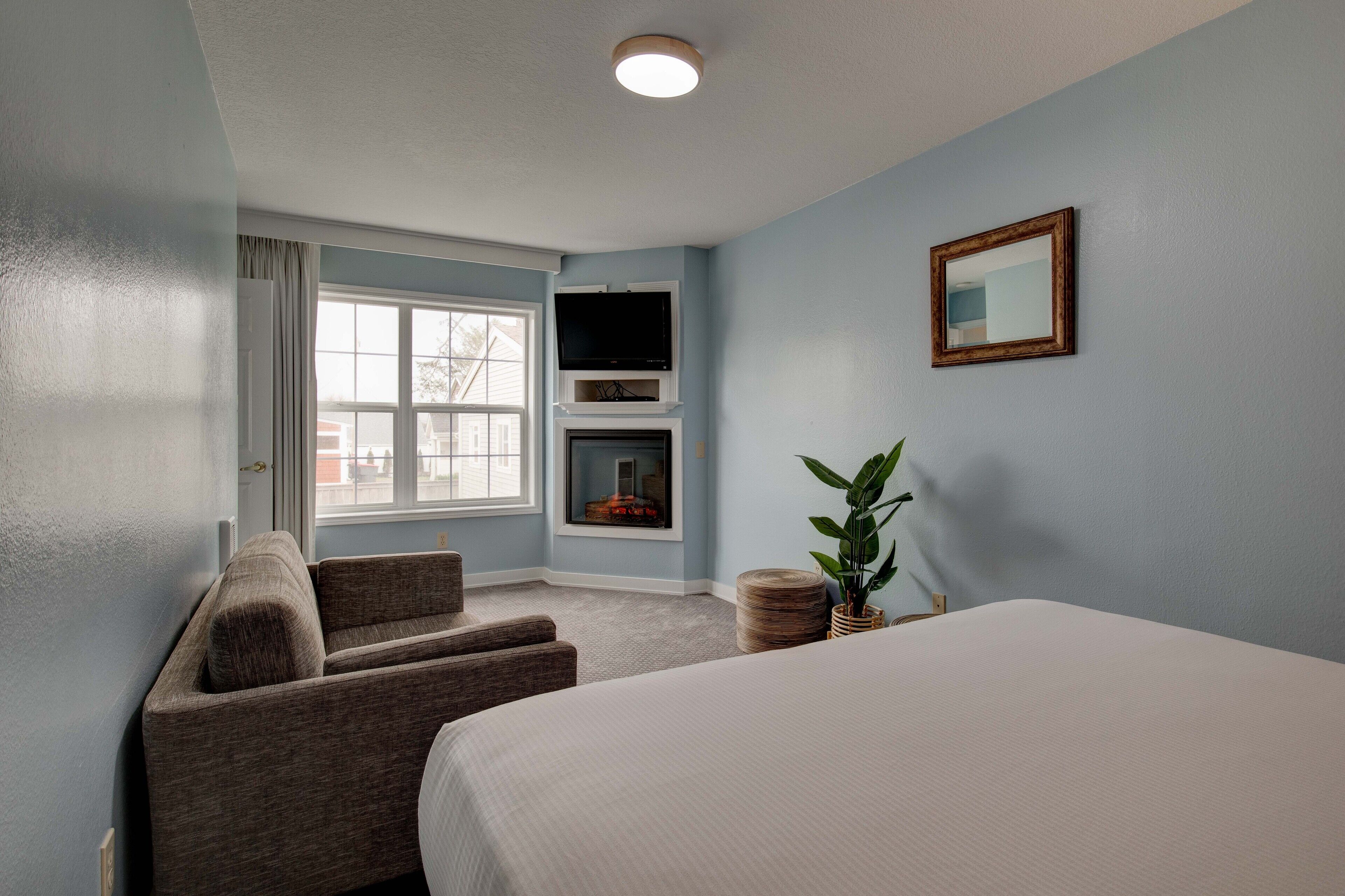 A hotel room with a bed, a seating area with a sofa and armchairs, a television mounted on the wall, a fireplace, and a window with curtains.