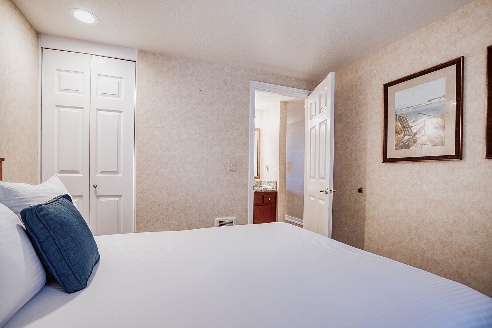 A bedroom with a bed, two doors, a framed picture, and a bathroom visible through an open door.
