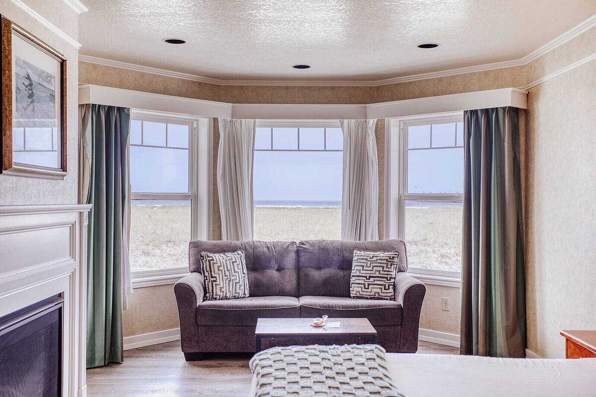 A room with a large window, a grey sofa with patterned cushions, a small table, and a woven ottoman.