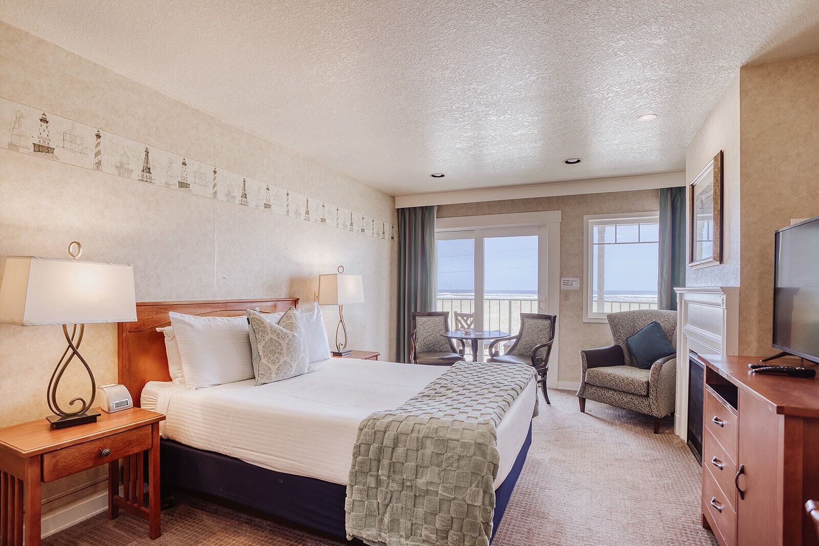 A hotel room with a large bed, a desk with a lamp, a TV, a chair, a balcony with a view, and a wall with a decorative pattern.