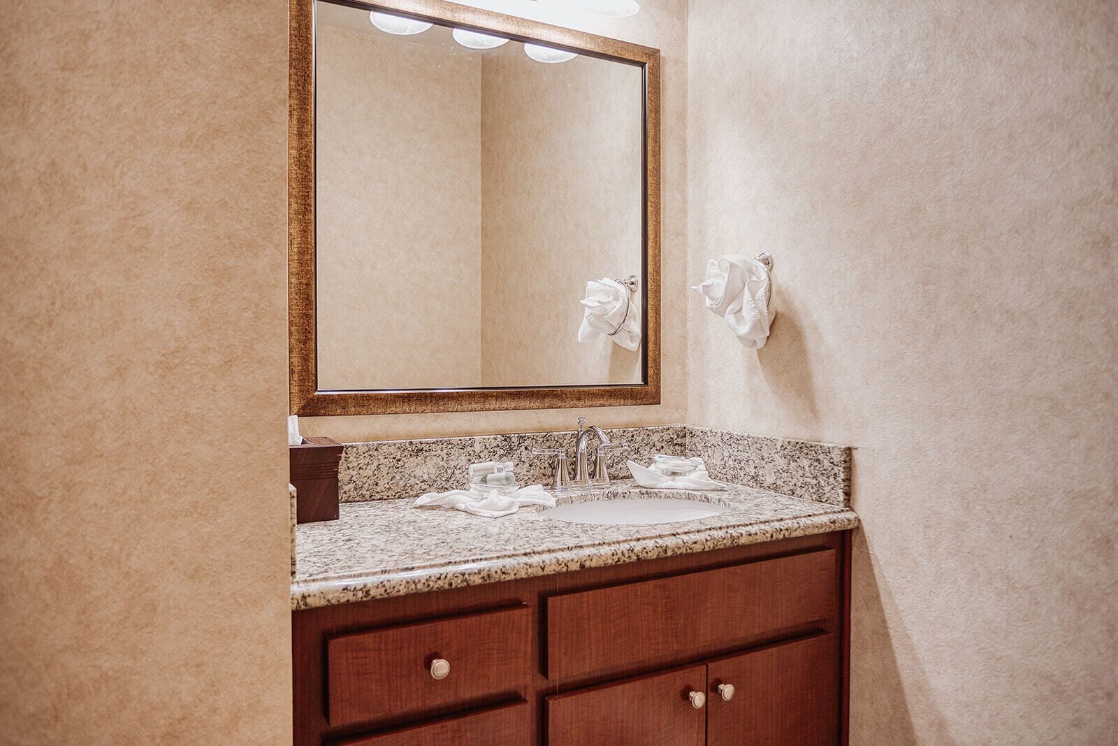 A bathroom with a granite countertop sink, a mirror with a wooden frame, and two towels hanging on the wall.