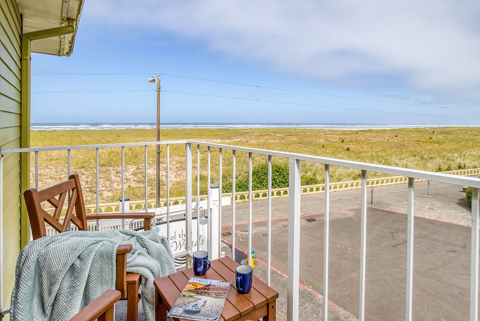 A balcony with a view of the beach, a wooden chair, a table with a magazine and two mugs, and a railing.