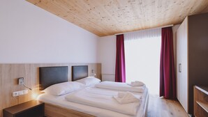 Ein modernes Hotelzimmer mit Holzboden, einem großen Bett mit weißen Leinen, einem Nachttisch mit Lampe und Blick ins Freie durch ein Fenster mit roten Vorhängen.