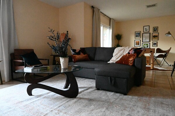 A living room with a dark grey sofa, a glass coffee table, and a potted plant. - Spacious Condo with game room, office and large kitchen. 5 min. to Sunrise Mall! (Brownsville)