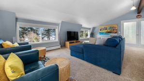 Ein geräumiges Wohnzimmer mit zwei blauen Sofas, einem Fernseher und großen Fenstern mit Blick auf Bäume.