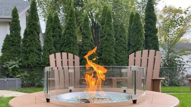 A round outdoor fire pit with a glass enclosure, surrounded by chairs, set in a grassy yard with trees in the background. - Sunny 3BR Craftsman Bungalow | Bay View (Milwaukee)