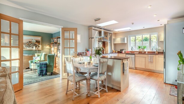 Une cuisine spacieuse avec un plancher en bois, un coin repas avec une table et des chaises, et une vue sur l’extérieur grâce à de grandes fenêtres.