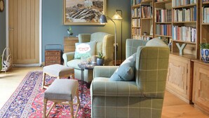 Une pièce avec un fauteuil vert, un pouf et une petite table sur laquelle se trouve une lampe. On y trouve également une bibliothèque remplie de livres et un tableau accroché au mur.