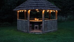 Un kiosque en bois avec un toit de chaume, des guirlandes lumineuses et une table centrale entourée de chaises.