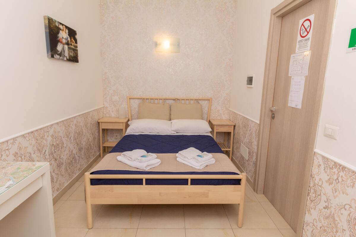 Ein ordentlich bezogenes Bett mit zwei Kissen und zwei gefalteten Handtüchern, ein Holzkopfteil, ein Nachttisch mit Lampe und eine Tür mit einem 'Nichtraucher'-Schild.