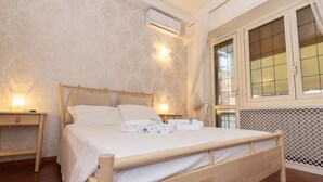 A neatly made bed with white linens and towels, a bedside table with a lamp, and an air conditioning unit on the wall. - Aurelio Deluxe Rooms (Rome)