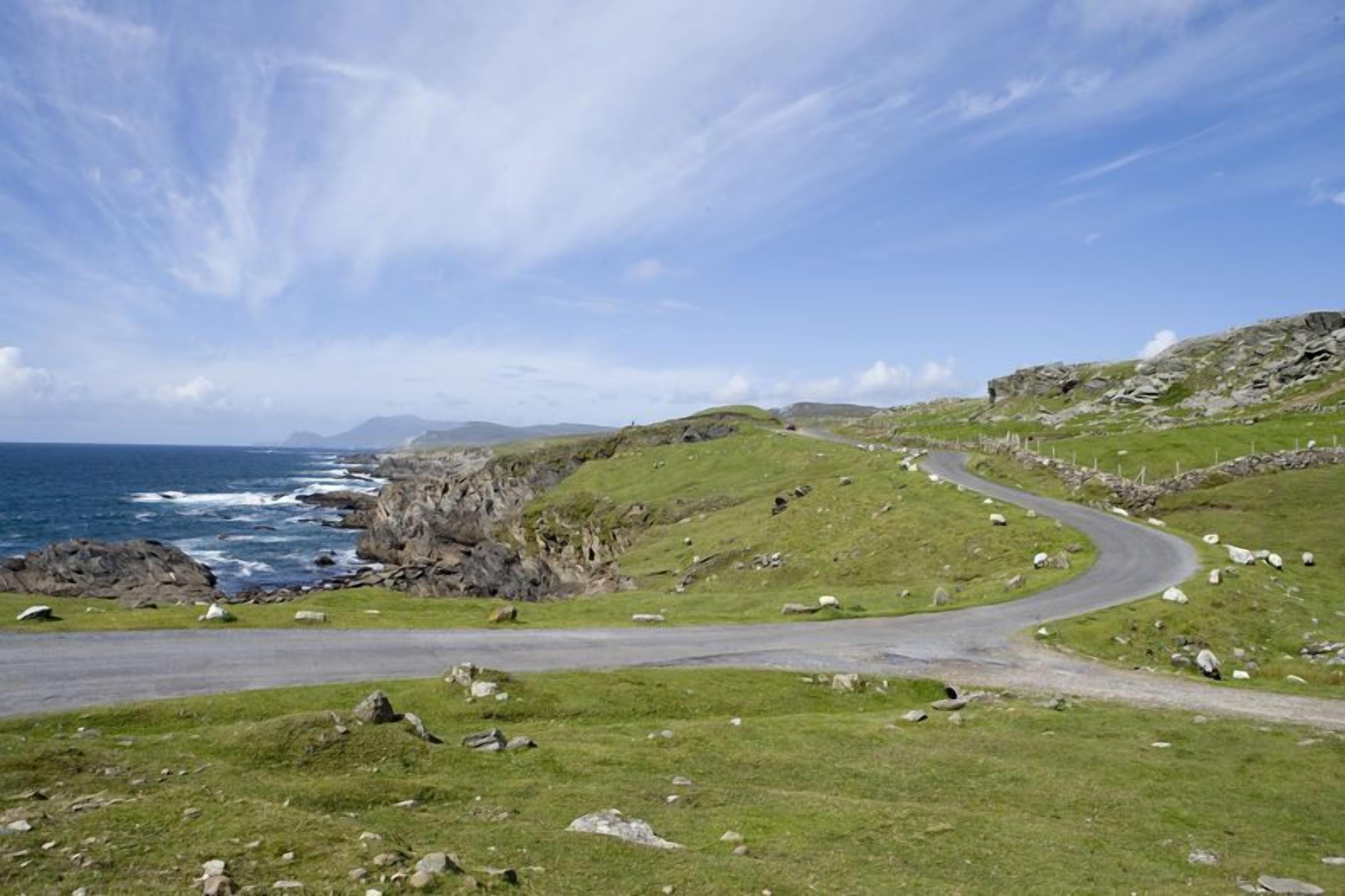 Una carretera costera serpentea a través de un paisaje herboso con afloramientos rocosos y el mar al fondo.