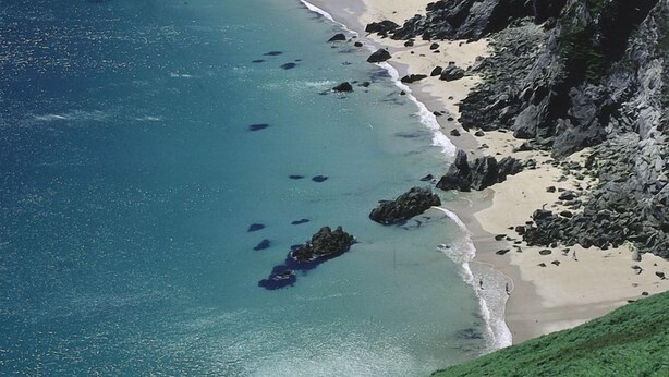 Vue aérienne d’une zone côtière avec une plage de sable, des affleurements rocheux et une eau d’un bleu limpide.