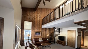A spacious living room with a high ceiling, a wooden wall, a staircase, a sofa, a coffee table, and a television.