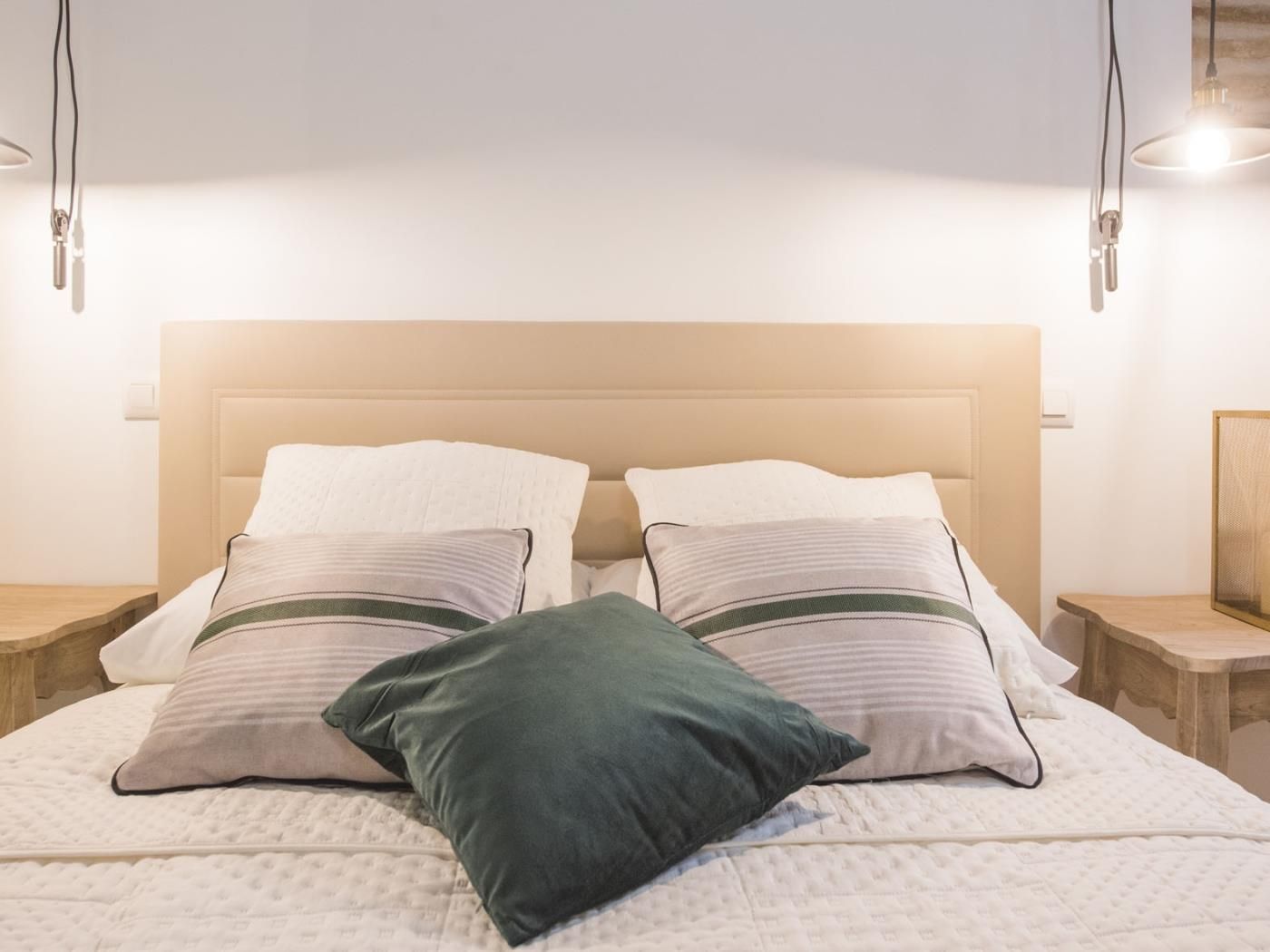 Una cama bien hecha con cabecera, dos mesitas de noche y una lámpara fijada a la pared.