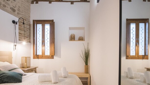 Una camera da letto con un letto, comodini, uno specchio, una finestra con persiane e una piccola mensola con oggetti decorativi.