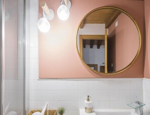 A bathroom with a round mirror, white tiles, a white bathtub, a small plant, and a soap dispenser.