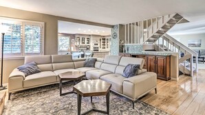 A spacious living room with a large L-shaped sectional sofa, a wooden coffee table, and a patterned rug. A kitchen with white cabinets and a tiled backsplash is visible in the background. - Near Shops & Eats! Centennial Home w/ Sunroom (Centennial)