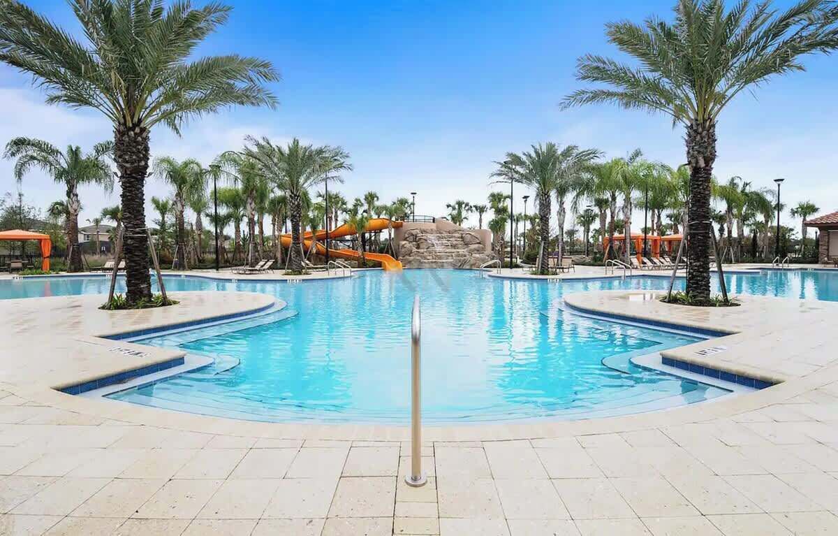 A large outdoor swimming pool with palm trees, a water slide, and a rock formation in the background.