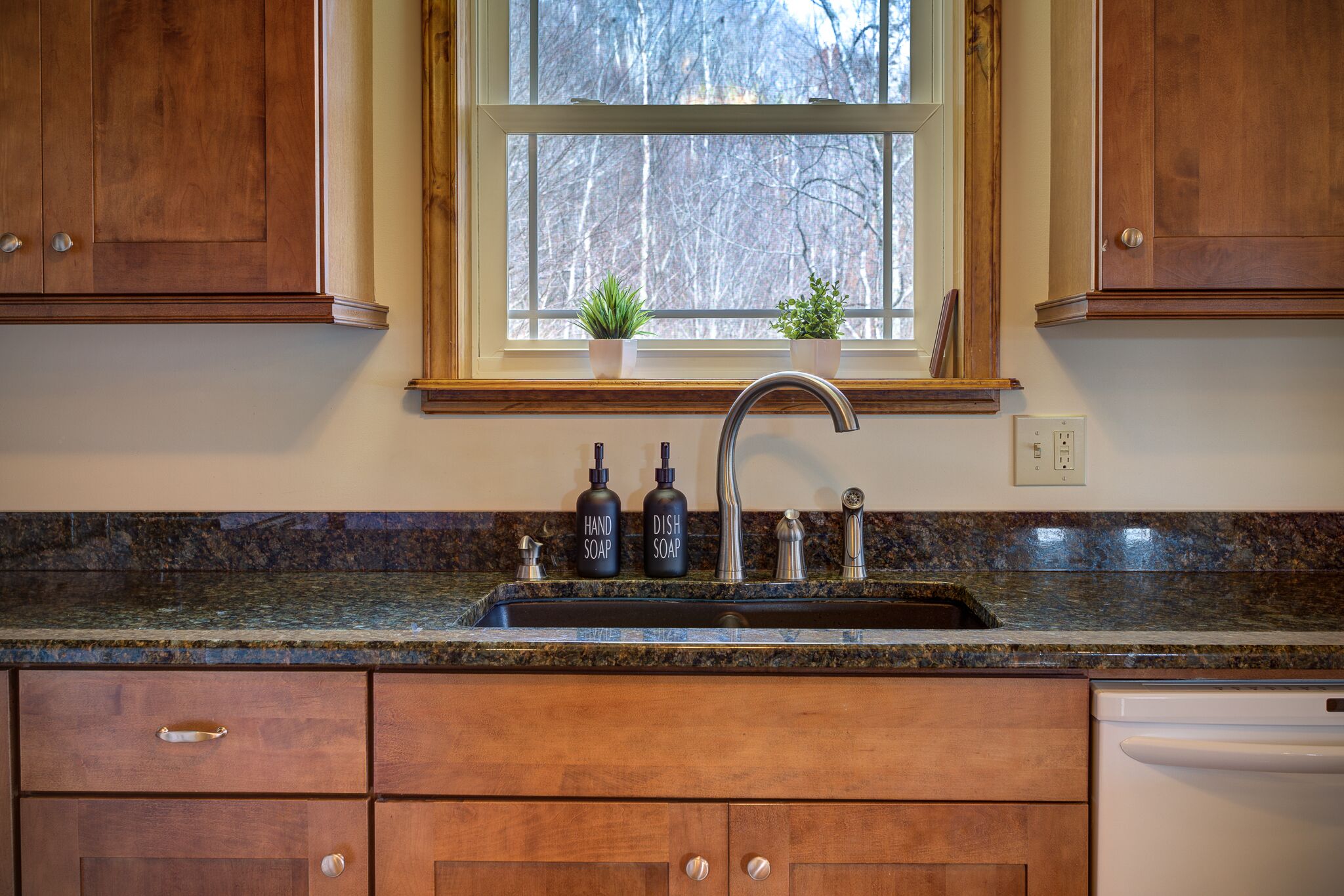 A kitchen with wooden cabinets, a granite countertop, a stainless steel sink, and two potted plants on the windowsill.