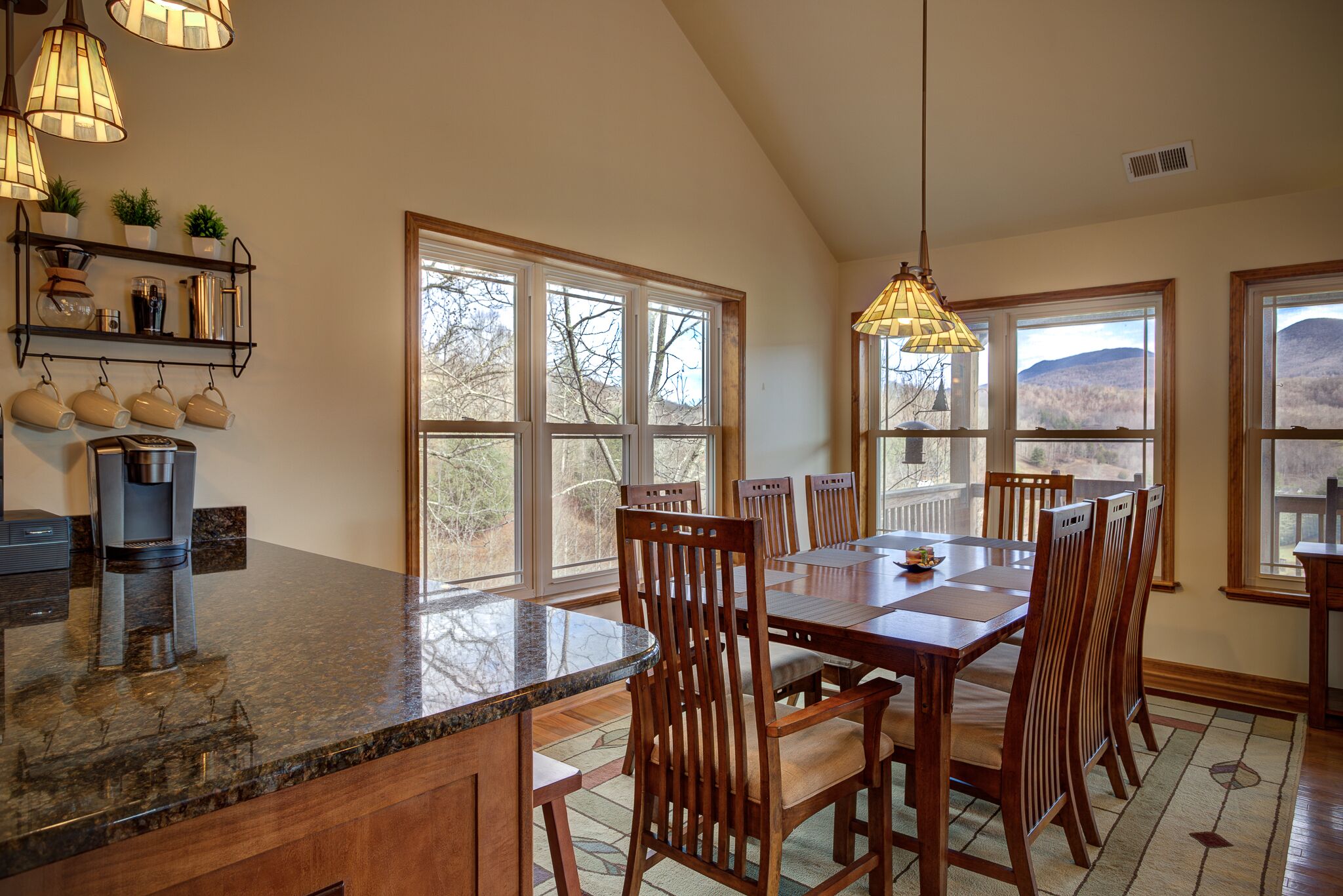 A kitchen with a large island, wooden chairs, and a dining table. There are multiple windows providing a view of the outdoors.