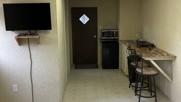 A compact living space with a flat-screen TV mounted on the wall, a small kitchenette with a microwave, and a dining area with two stools.