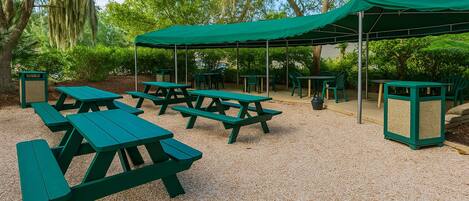 Un espace extérieur aménagé avec des tables et des bancs de pique-nique verts, entouré d’arbres et recouvert d’une pergola verdoyante.