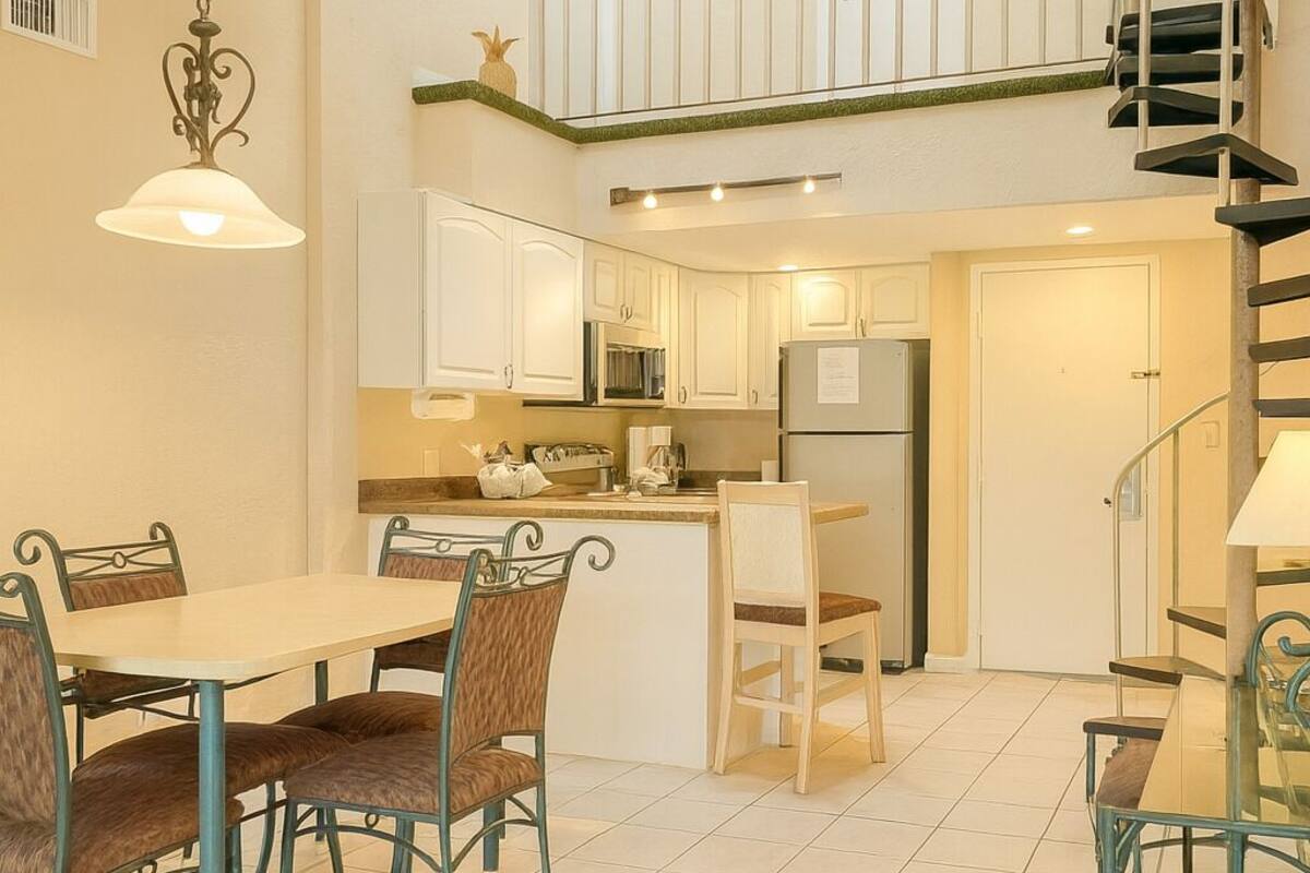Una cocina moderna con armarios blancos, una zona de comedor con mesa y sillas, y una escalera que sube a un nivel superior.