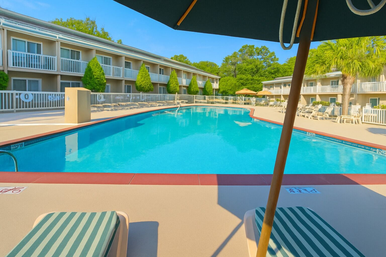 A swimming pool with lounge chairs and umbrellas, surrounded by a fence and a building with balconies.