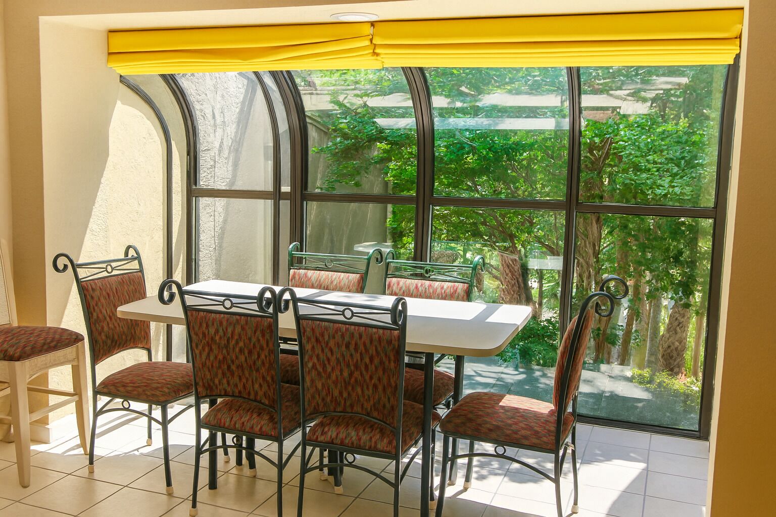 Zona pranzo con tavolo e sedie, ampie finestre con vista su piante e un tendaggio giallo.