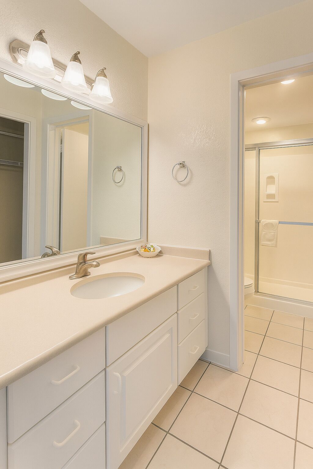 A bathroom with a sink, mirror, and three lights above it. There are two towel rings on the wall and a shower door visible in the background.