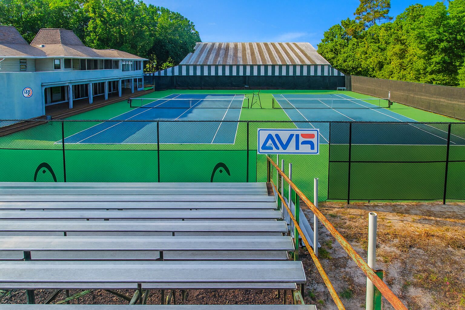 Una cancha de tenis con una red, rodeada por una cerca, y un edificio con un letrero que dice 'AVR' al fondo.
