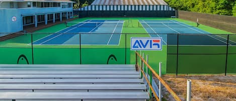 Una cancha de tenis con una red, rodeada por una cerca, y un edificio con un letrero que dice 'AVR' al fondo.