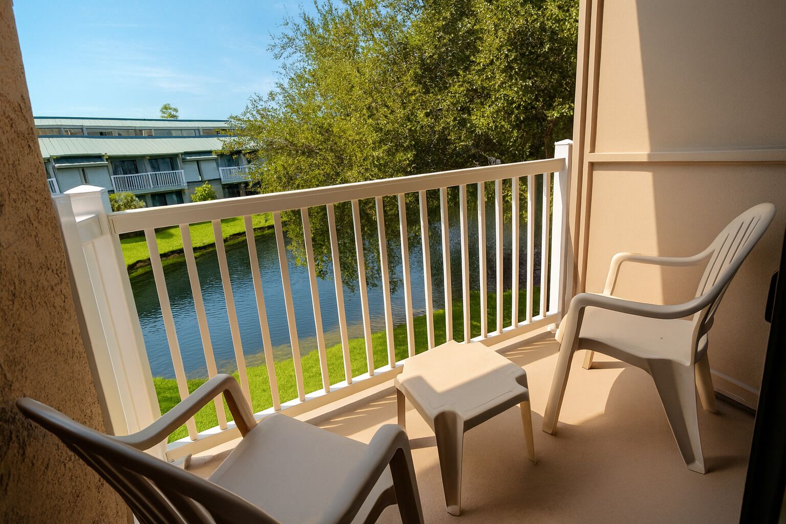 Un balcon avec des balustrades blanches, deux chaises beiges et une petite table. On aperçoit une étendue d’eau et de la verdure à l’arrière-plan.