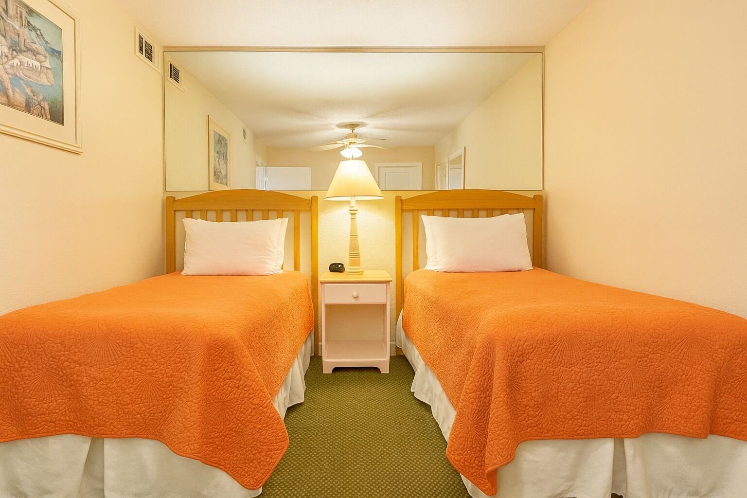 A hotel room with two single beds, each with an orange bedspread, a nightstand with a lamp, and a green carpet.